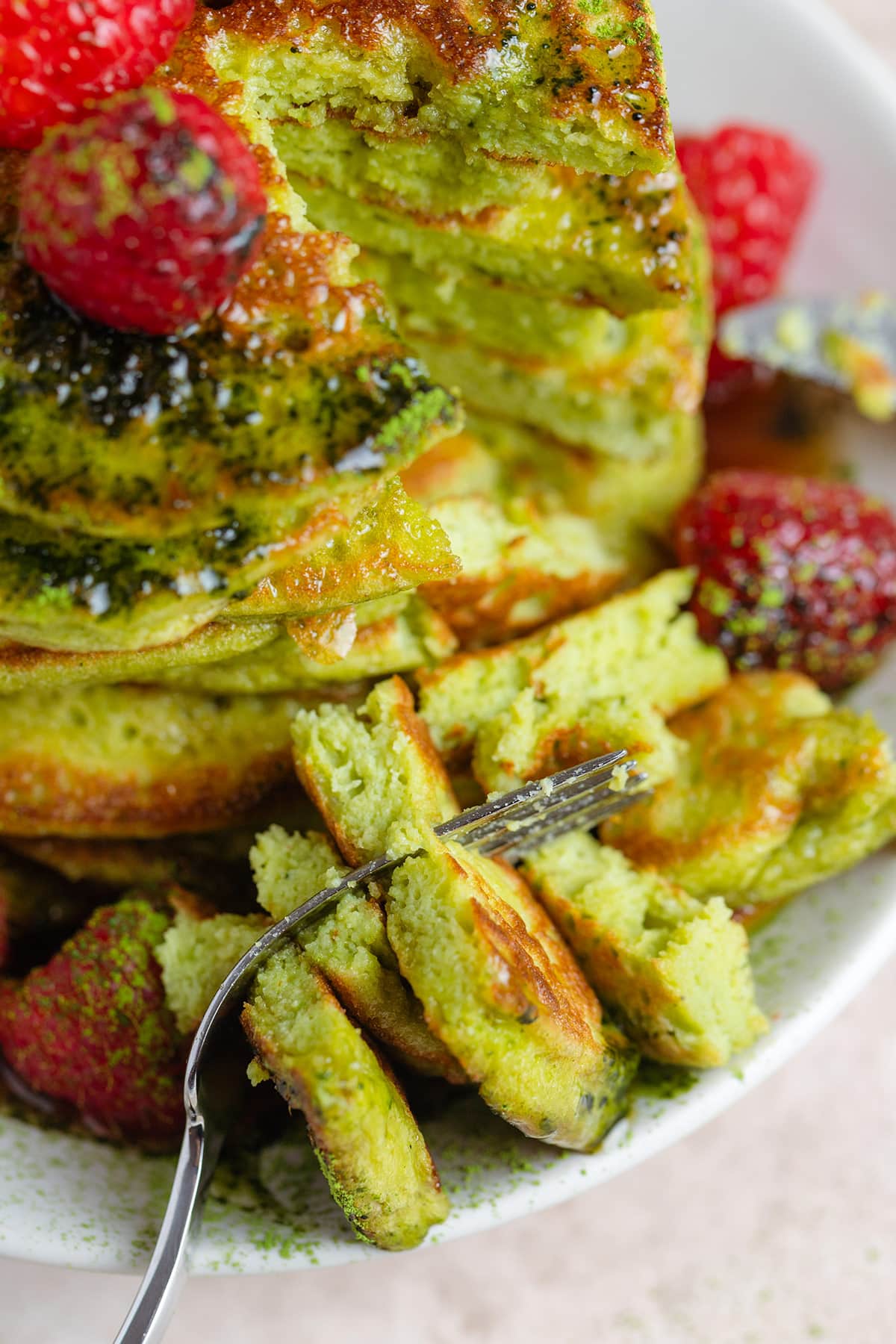 A stack of green matcha pancakes with raspberries drizzled with maple syrup and dusted with matcha powder with a fork taking out a bite.