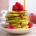 A stack of green matcha pancakes with fresh raspberries being drizzled with maple syrup on a white plate.