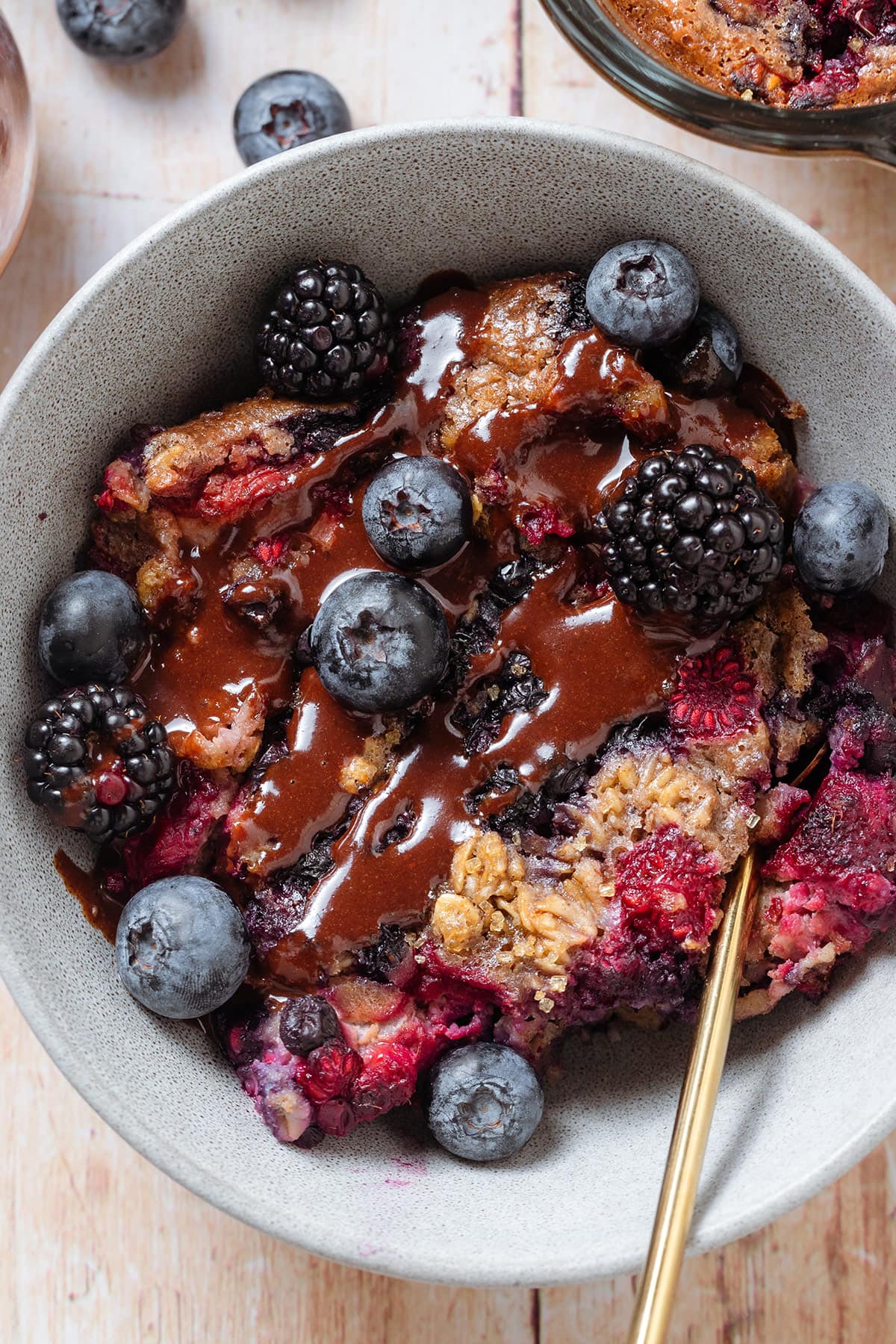 Baked berry oatmeal in a grey bowl topped with blueberries and blackberries and drizzled with chocolate sauce.