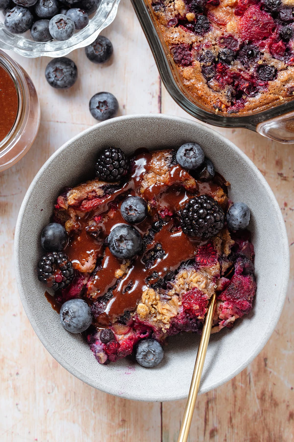 Baked berry oatmeal in a grey bowl topped with blueberries and blackberries and drizzled with chocolate sauce.