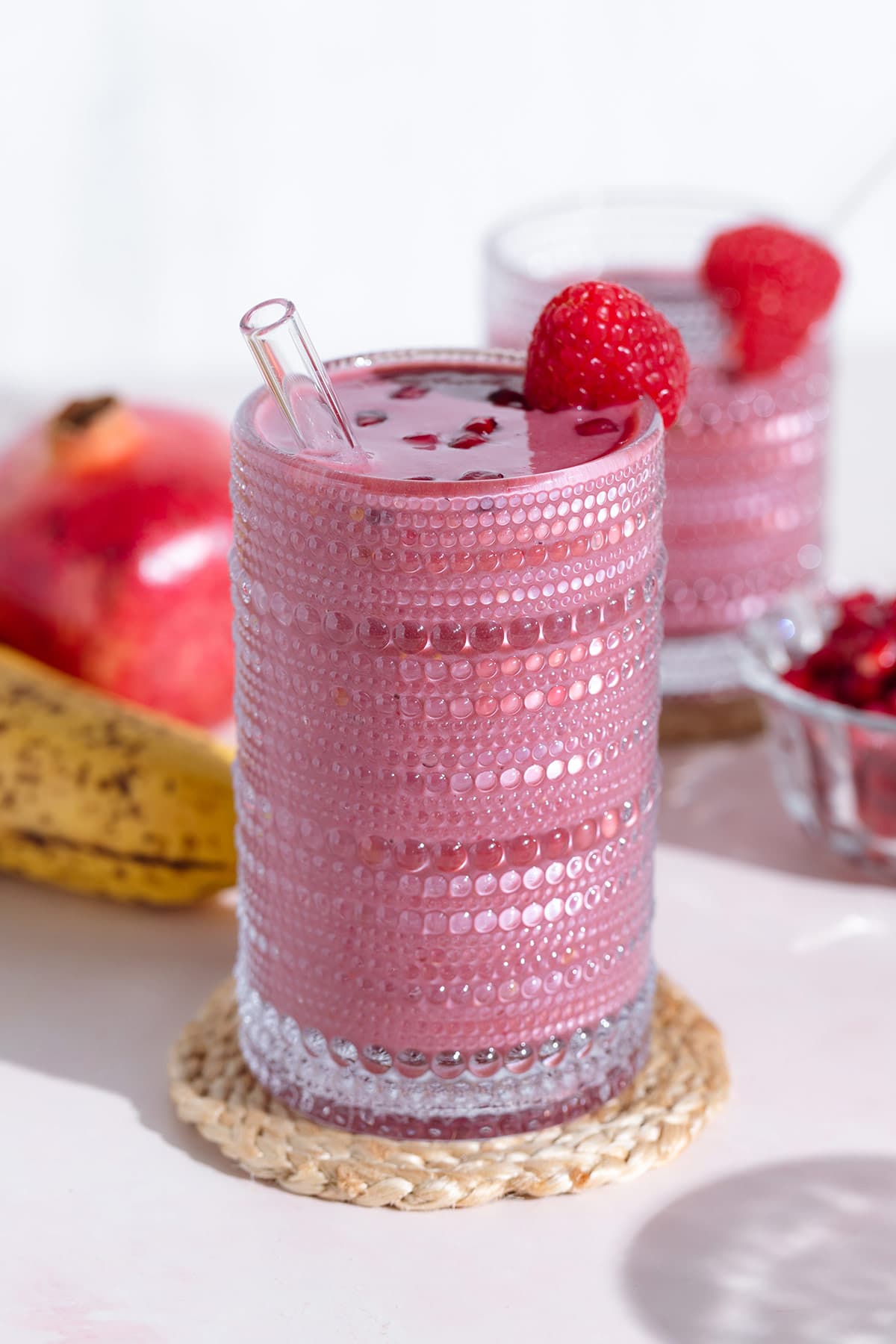 A pink smoothie in a tall glass garnished with pomegranate seeds and a raspberry on a white background.