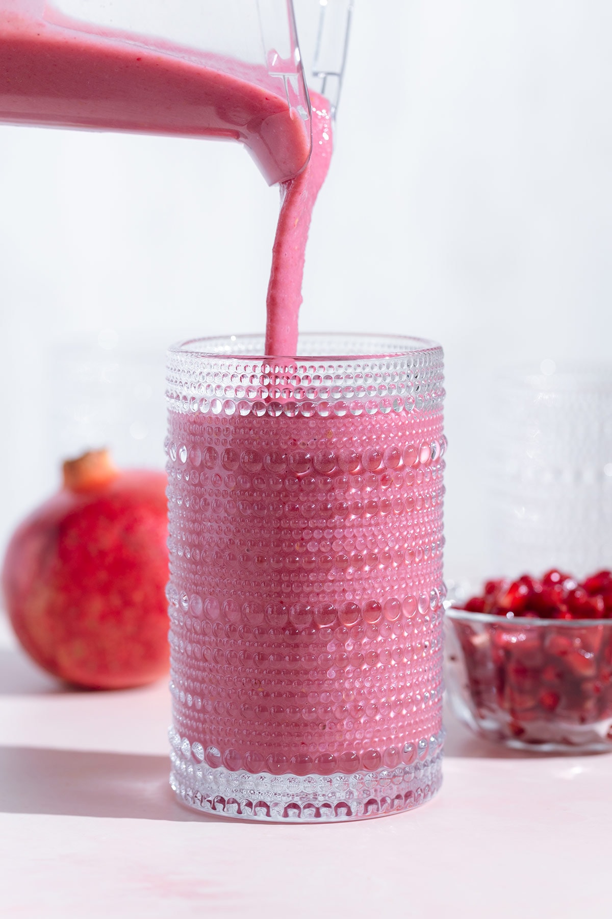Pink smoothie being poured into a tall glass on a white and pink background.