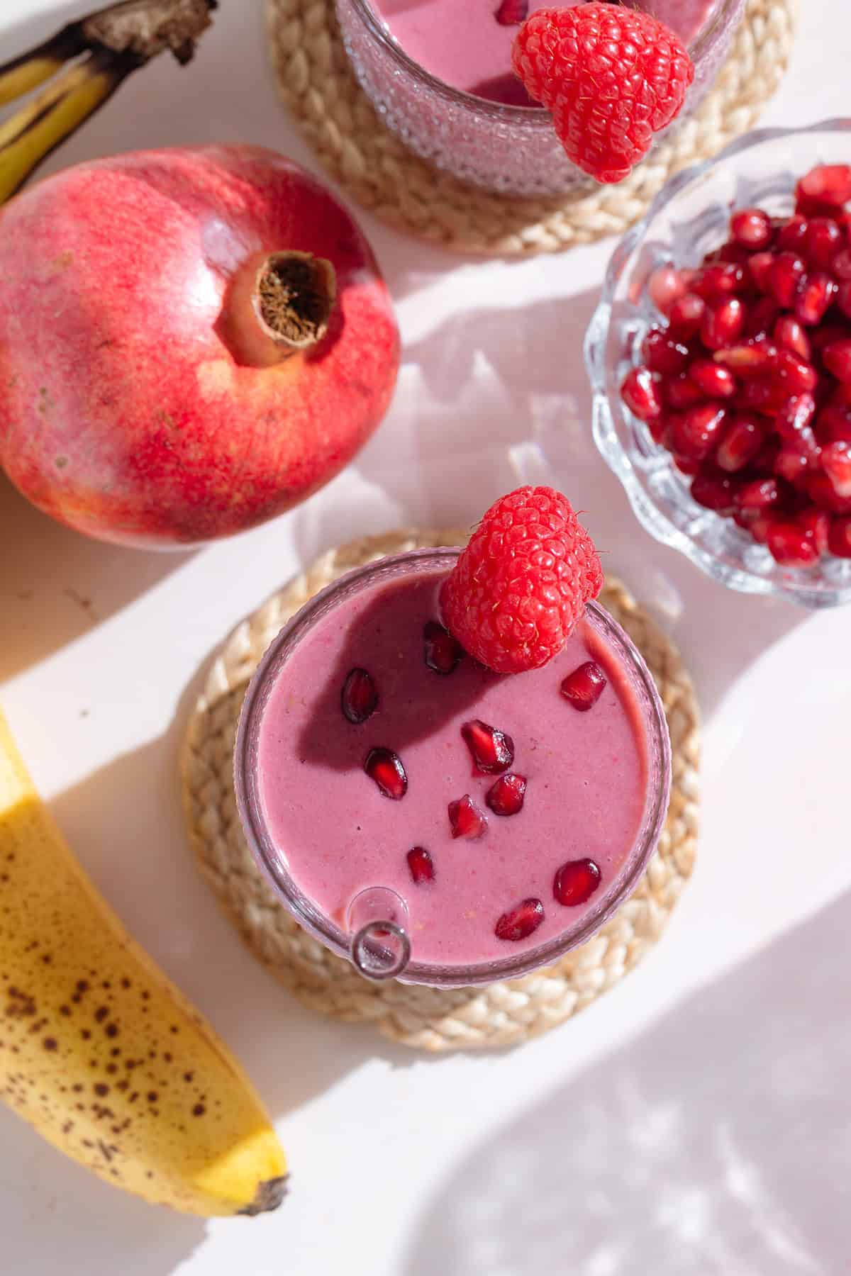 A pink smoothie in a tall glass garnished with pomegranate seeds and a raspberry on a white background.