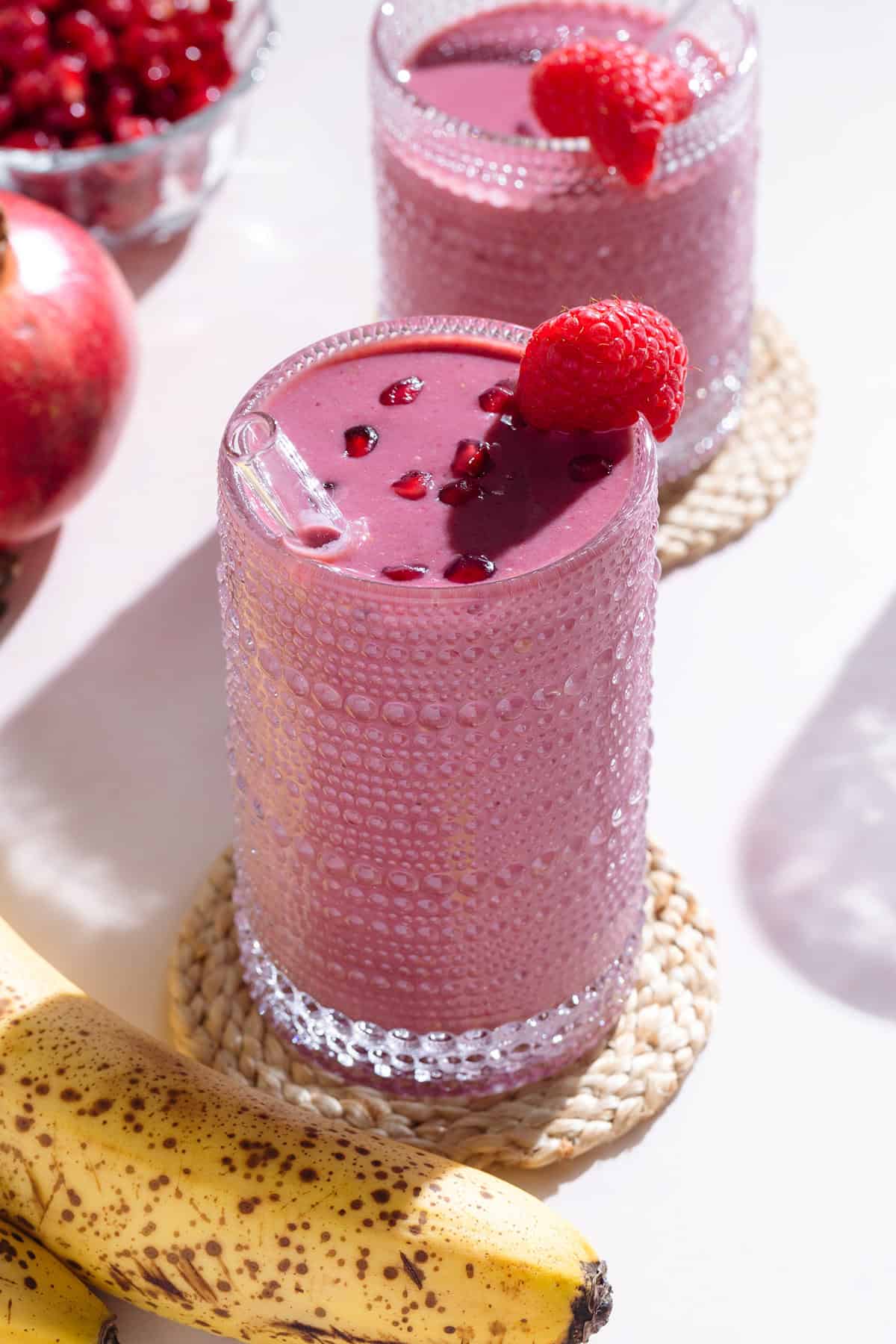 A pink smoothie in a tall glass garnished with pomegranate seeds and a raspberry on a white background.