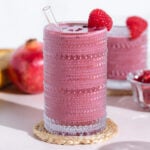 A pink smoothie in a tall glass garnished with pomegranate seeds and a raspberry on a white background.