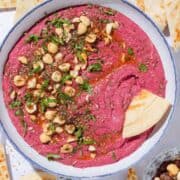 Bright pink beet dip in a white bowl with a blue rim garnished with hazelnuts, parsley, and olive oil with a naan triangle dipped into the dip.