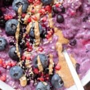 Bright purple blueberry oatmeal in a white bowl garnished with blueberries, nut butter, and hemp seeds on a dark wooden background.