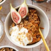 A slice of carrot cake baked oatmeal in a white bowl garnished with yogurt and fresh figs on a wooden background.