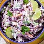 Colorful cilantro lime slaw with purple cabbage in a blue bowl garnished with lime slices on a yellow plate.