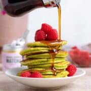 A stack of green matcha pancakes with fresh raspberries being drizzled with maple syrup on a white plate.
