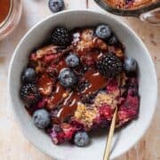 Baked berry oatmeal in a grey bowl topped with blueberries and blackberries and drizzled with chocolate sauce.