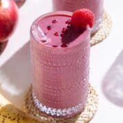A pink smoothie in a tall glass garnished with pomegranate seeds and a raspberry on a white background.