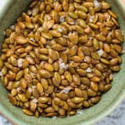 Roasted pepitas in a small green ceramic bowl sprinkled with flaky sea salt.