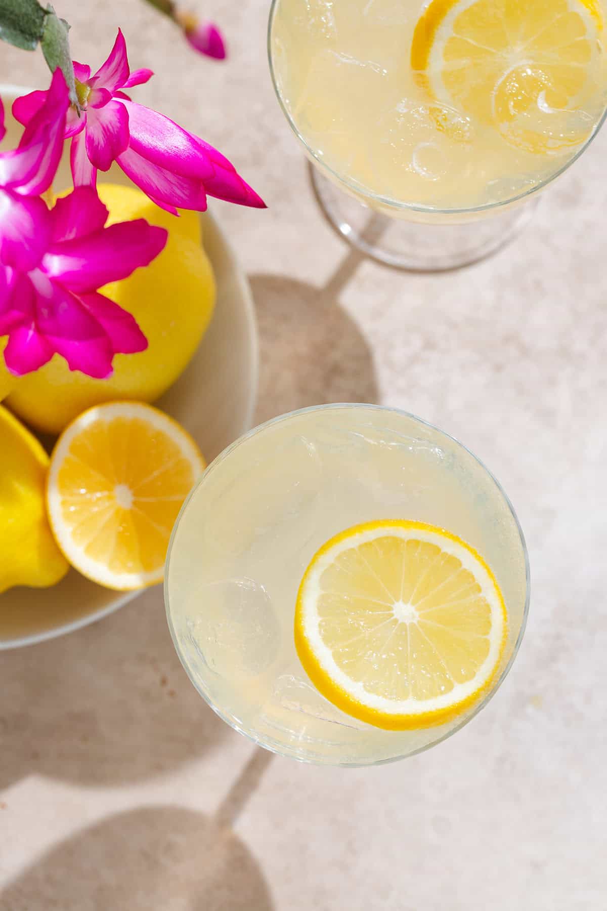 Yellow lemon margarita in a coupe glass with ice and a lemon slice as garnish on beige background with lemons and purple flowers in the background.