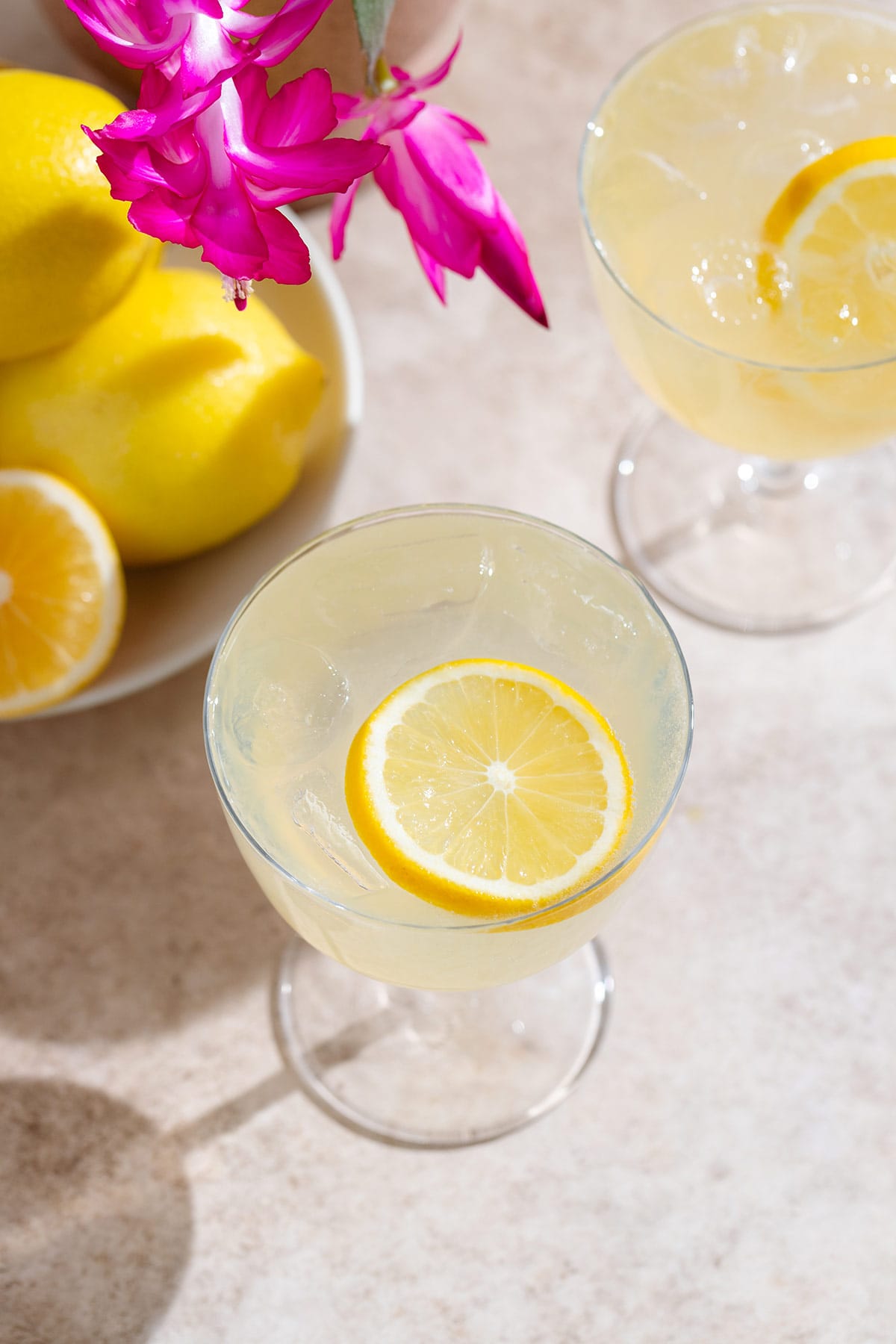 Yellow lemon margarita in a coupe glass with ice and a lemon slice as garnish on beige background with lemons and purple flowers in the background.