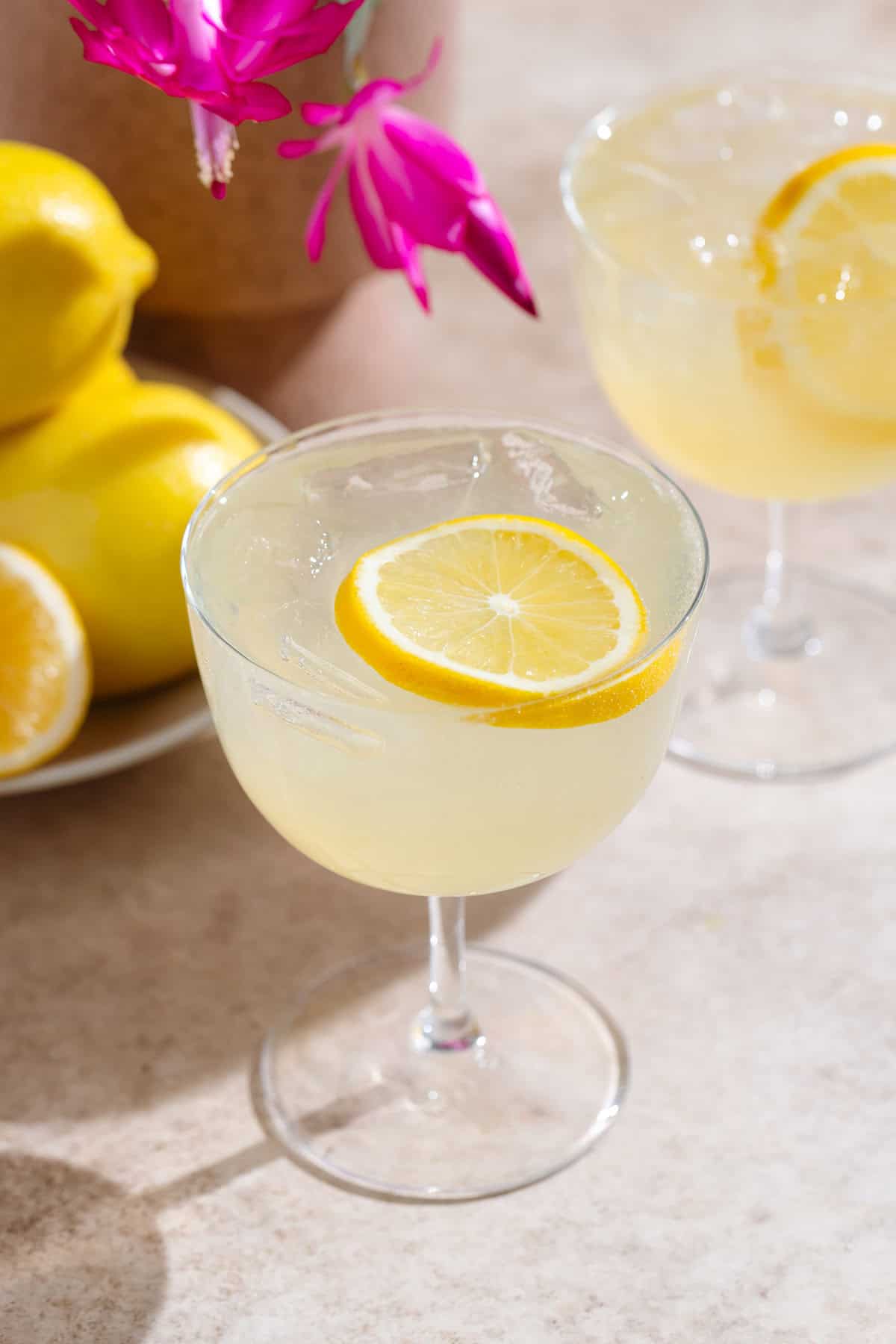 Yellow lemon margarita in a coupe glass with ice and a lemon slice as garnish on beige background with lemons and purple flowers in the background.