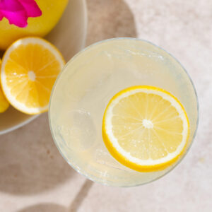 Yellow lemon margarita in a coupe glass with ice and a lemon slice as garnish on beige background with lemons and purple flowers in the background.