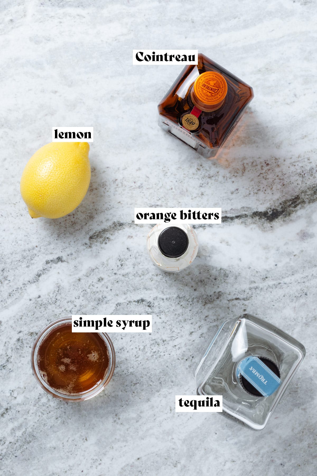Cointreau, tequila, lemon, orange bitters, and simple syrup laid out on a stone background.