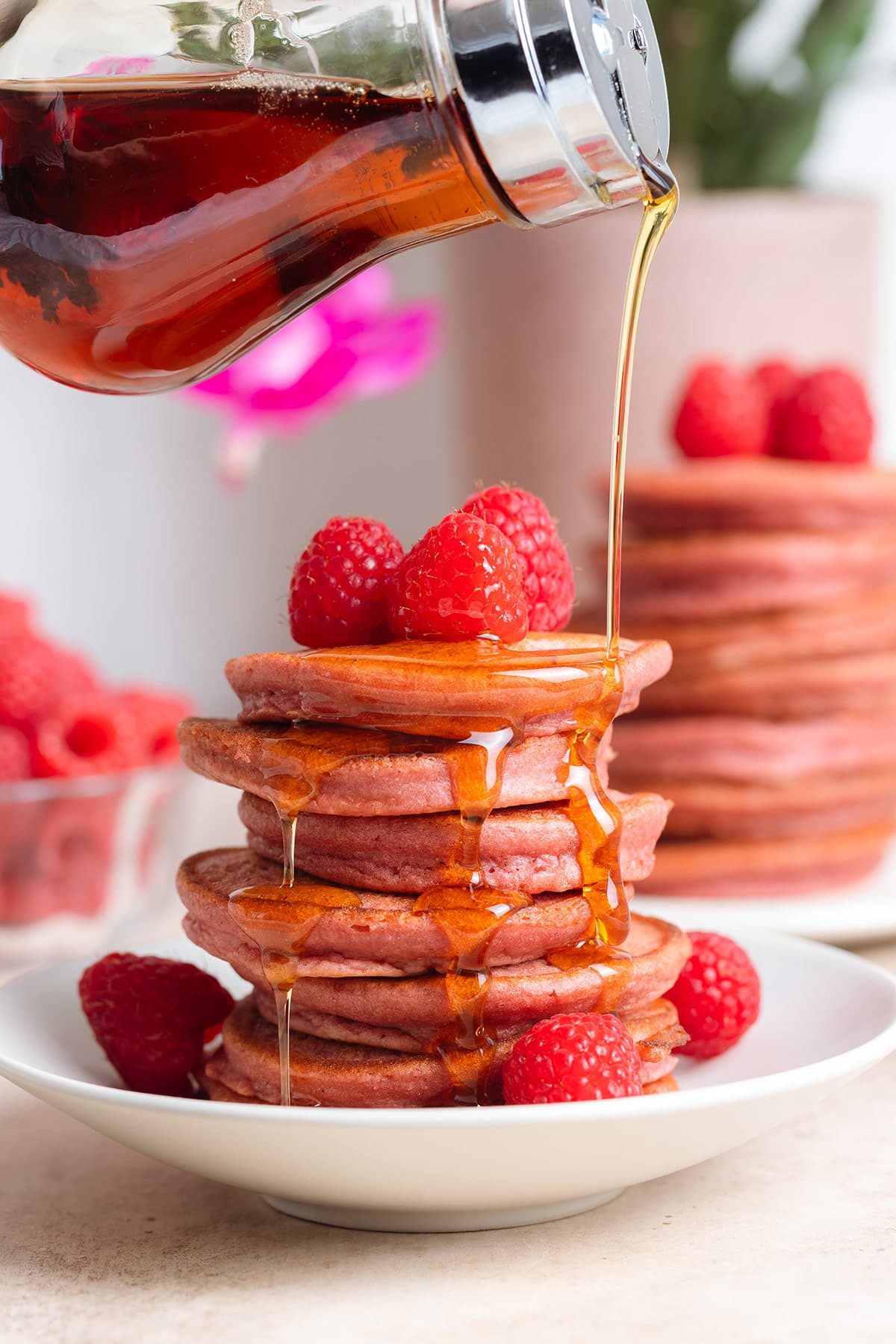 A stack of pink pancakes on a small white plate garnished with fresh raspberries and being drizzled with maple syrup.