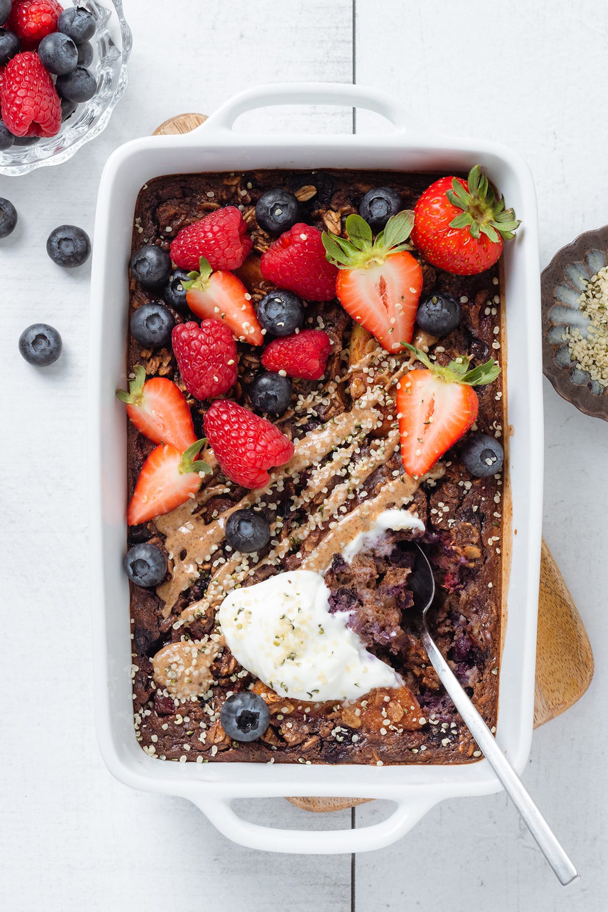 Baked oatmeal in a white baking dish garnished with fresh berries and almond butter with a spoon inserted on the right.