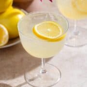 Yellow lemon margarita in a coupe glass with ice and a lemon slice as garnish on beige background with lemons and purple flowers in the background.