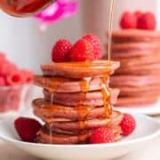 A stack of pink pancakes on a small white plate garnished with fresh raspberries and being drizzled with maple syrup.