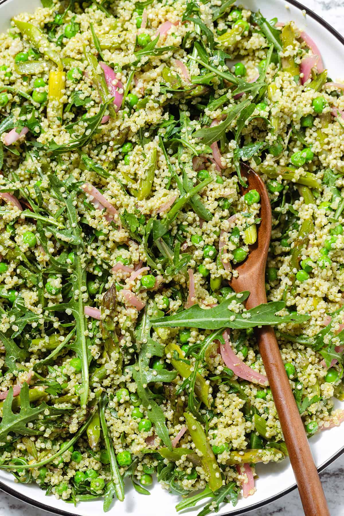 Quinoa salad with arugula, asparagus, and peas on a large serving platter with wooden spoon inserted in it.