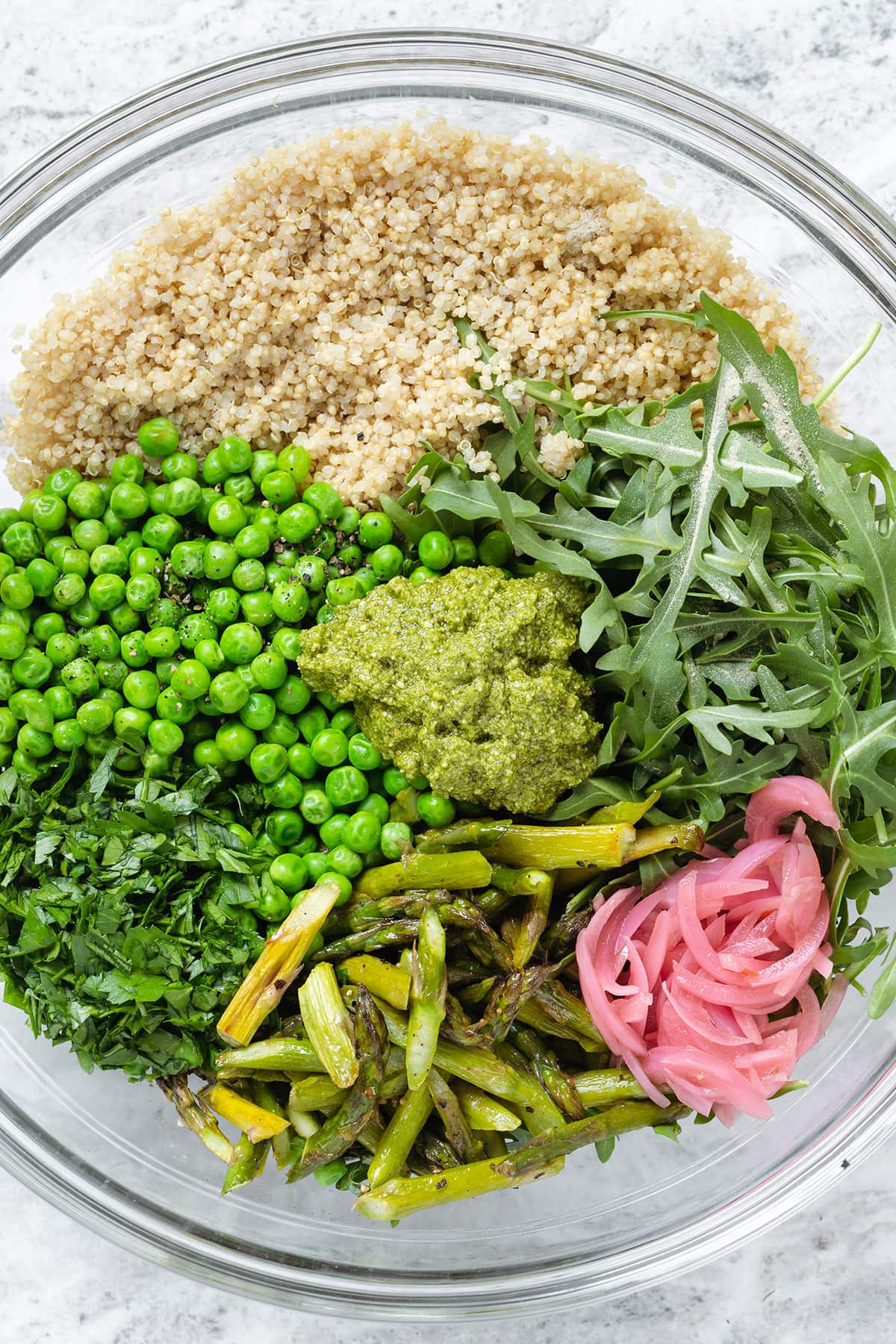 A large glass bowl with quinoa, arugula, roasted asparagus, peas, and red onion.