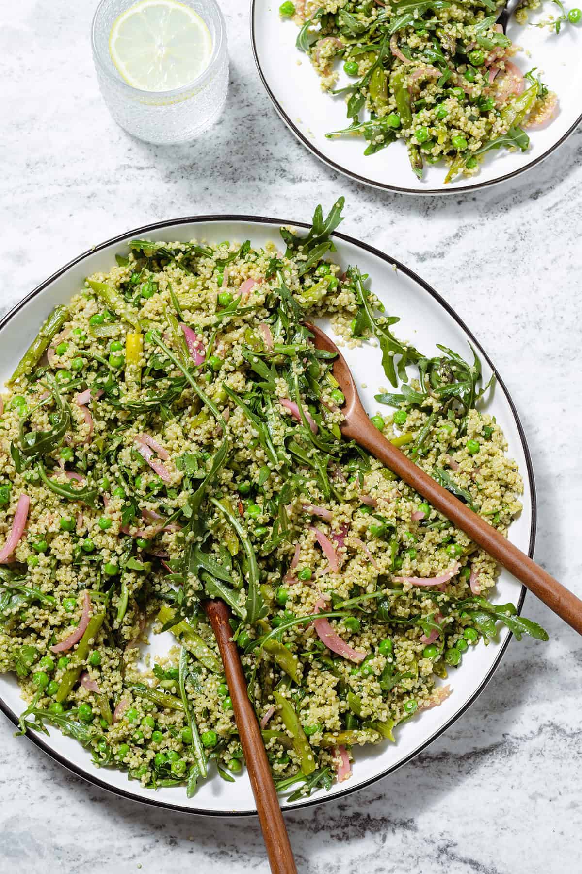 Quinoa salad with arugula, asparagus, and peas on a large serving platter with wooden spoons inserted in it.