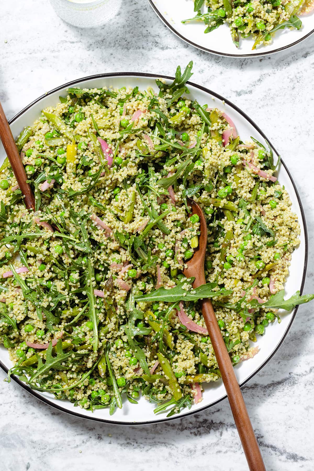 Quinoa salad with arugula, asparagus, and peas on a large serving platter with wooden spoons inserted in it.