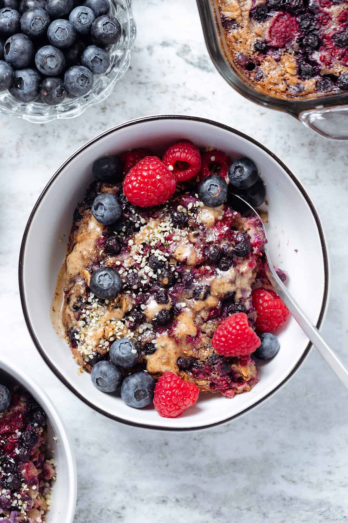 A slice of blueberry baked oatmeal in a white bowl with black rim garnished with berries and almond butter.