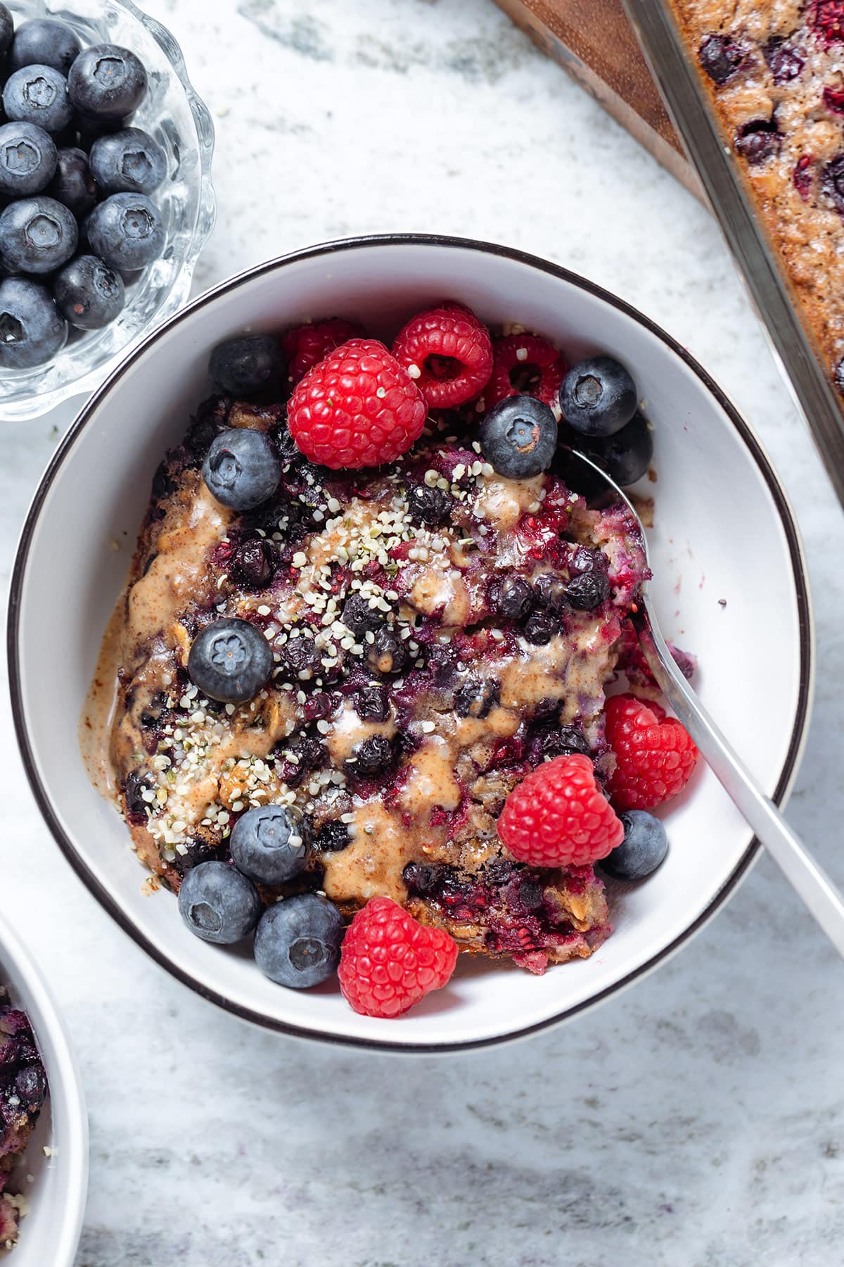 A slice of blueberry baked oatmeal in a white bowl with black rim garnished with berries and almond butter.