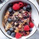 A slice of blueberry baked oatmeal in a white bowl with black rim garnished with berries and almond butter.