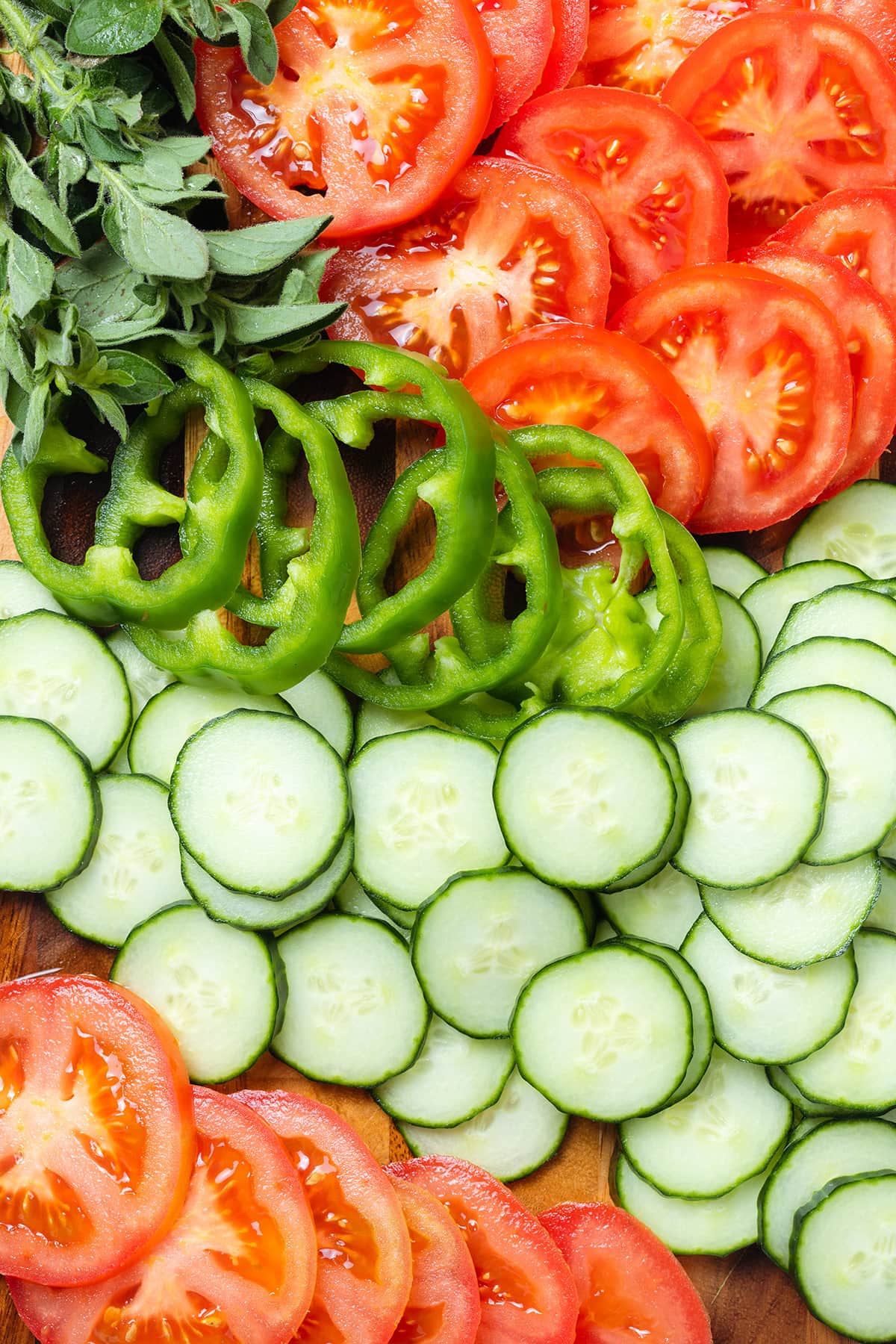 Sliced cucumbers, tomatoes, green bell peppers, and fresh oregano on a wooden cutting board.