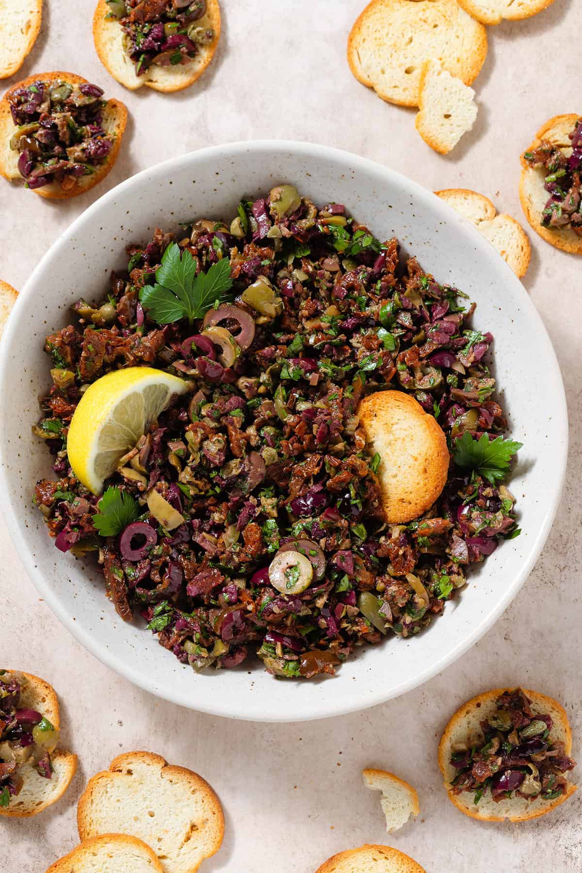 Olive tapenade in a beige bowl garnished with a lemon wedge and with crackers around the bowl.