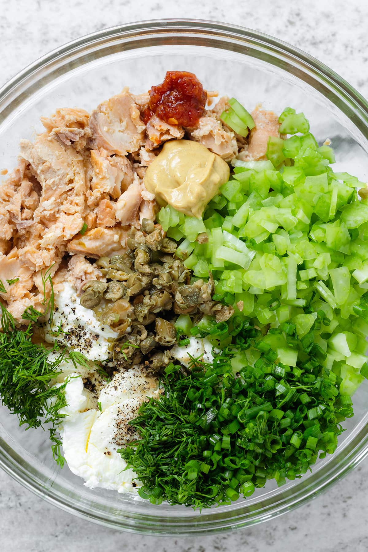 Canned salmon, chopped celery, herbs, and capers in a large glass bowl.