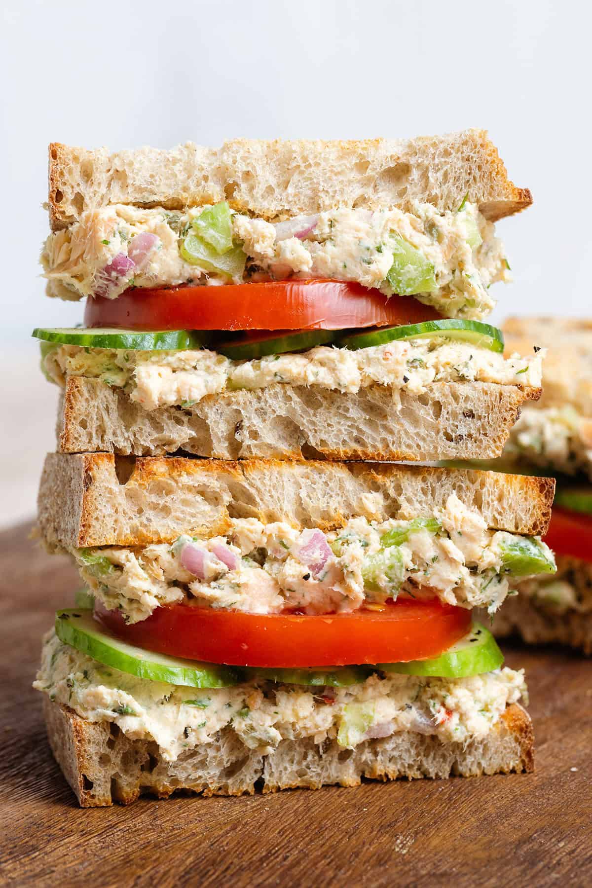 A sourdough sandwich stack with canned salmon spread, tomatoes, and cucumber on a wooden cutting board.