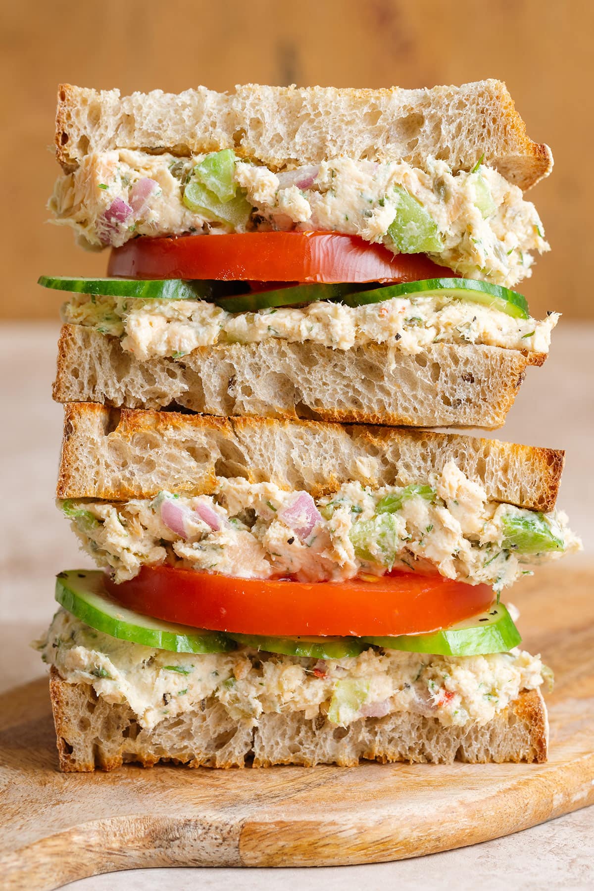 A sourdough sandwich stack with canned salmon spread, tomatoes, and cucumber on a wooden cutting board.