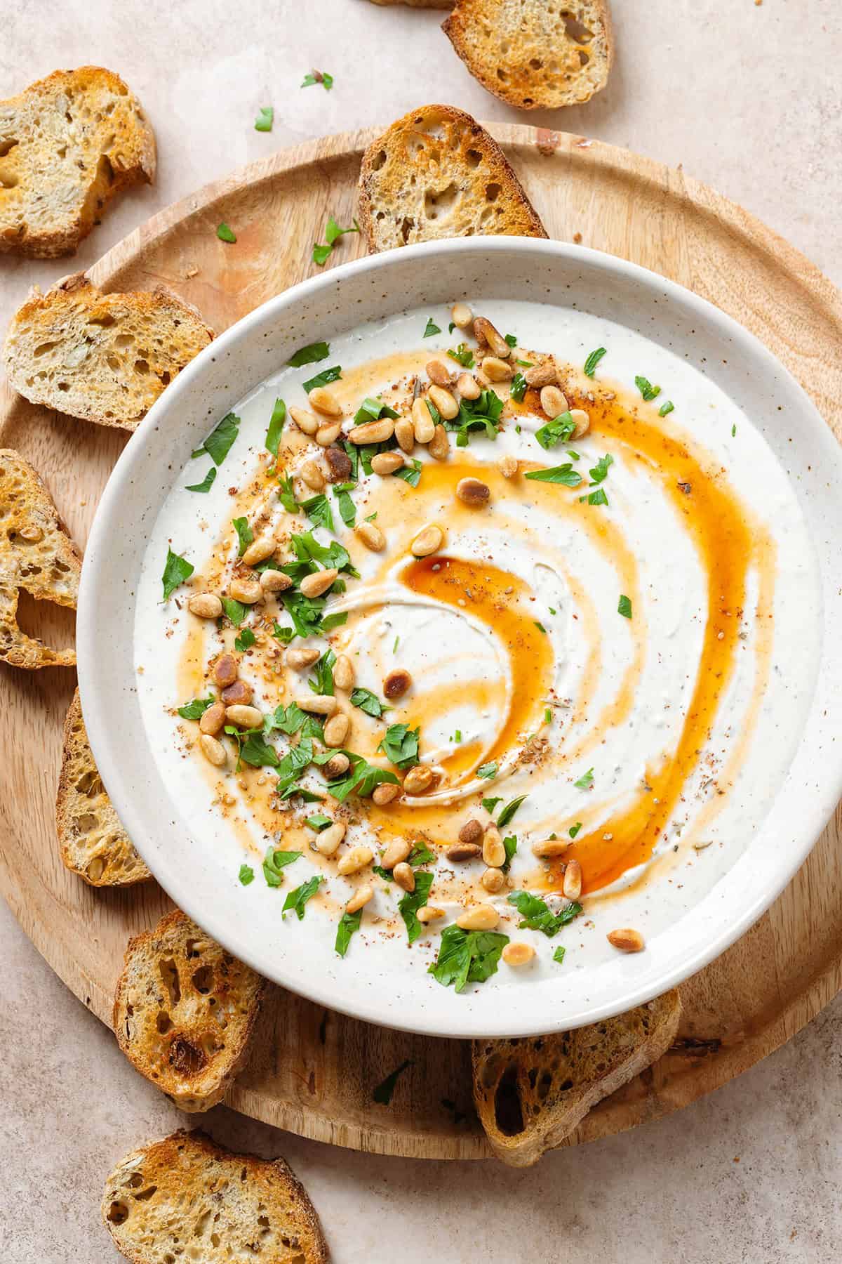 Whipped feta dup in a white bowl drizzled with hot honey and garnished with toasted pine nuts and fresh herbs.