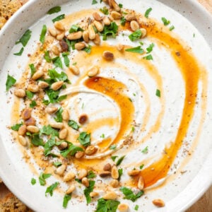 Whipped feta dup in a white bowl drizzled with hot honey and garnished with toasted pine nuts and fresh herbs.