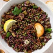 Olive tapenade in a beige bowl garnished with a lemon wedge and with crackers around the bowl.