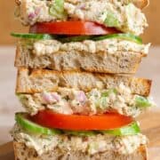 A sourdough sandwich stack with canned salmon spread, tomatoes, and cucumber on a wooden cutting board.