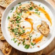 Whipped feta dup in a white bowl drizzled with hot honey and garnished with toasted pine nuts and fresh herbs.