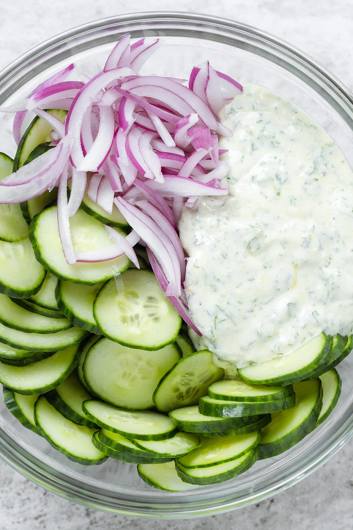 Sliced cucumber and red onion in a glass bowl with tzatziki dressing before mixing together.