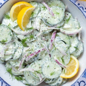 Creamy cucumber salad with red onion garnished with lemon wedges in a bowl with a blue rim.
