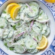 Creamy cucumber salad with red onion garnished with lemon wedges in a bowl with a blue rim.