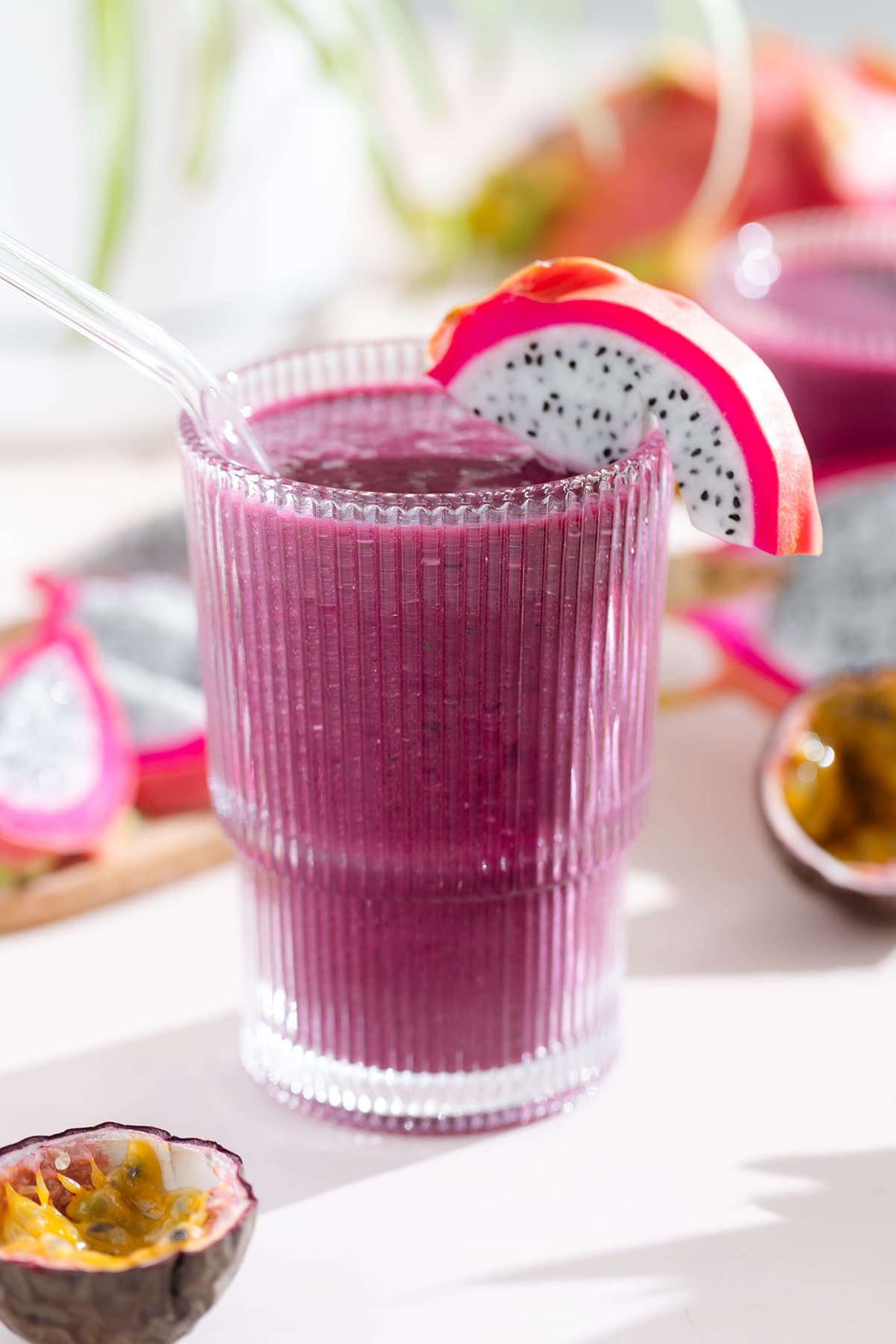 Bright purple smoothie in a tall glass with a glass straw garnished with white dragon fruit with more dragon fruit and passion fruit around the glass.