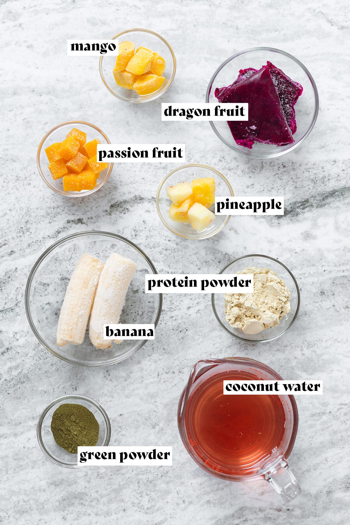 Ingredients like coconut water, frozen banana, dragon fruit, and mango on a grey stone background.
