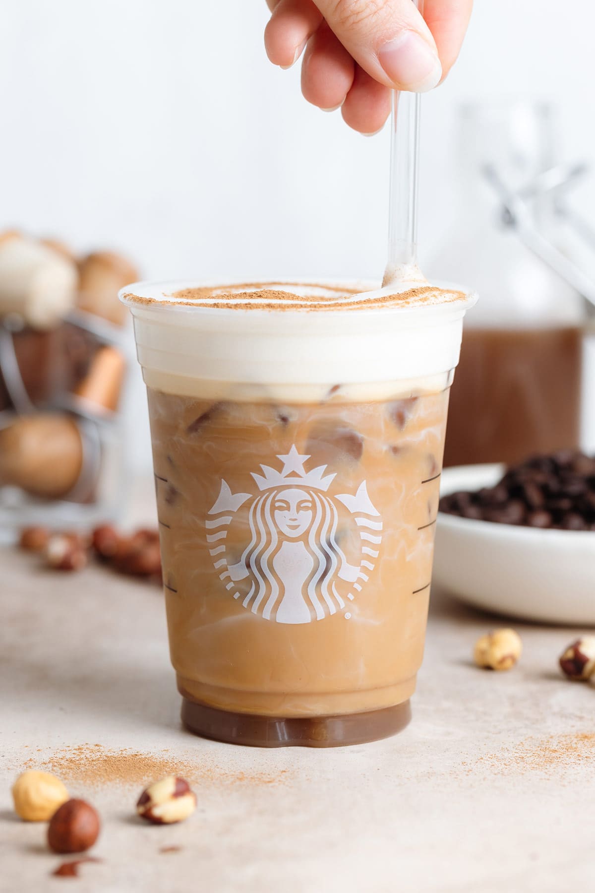 A plastic Starbucks cup with iced latte with frothy milk and a sprinkle of cinnamon with a hand stirring it using a glass straw.