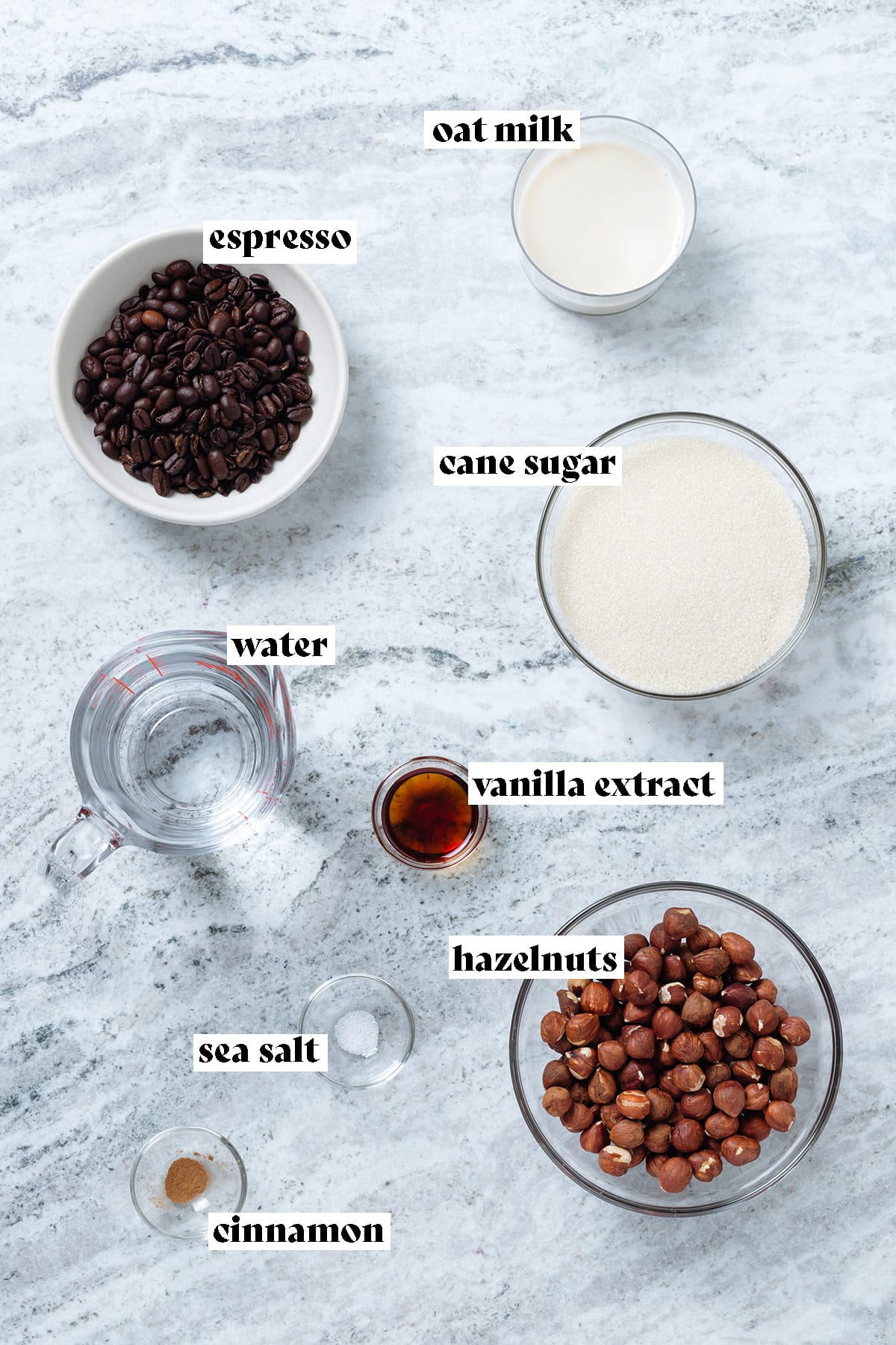 Ingredients for iced hazelnut latte like hazelnuts, coffee beans, and milk laid out on a stone background.